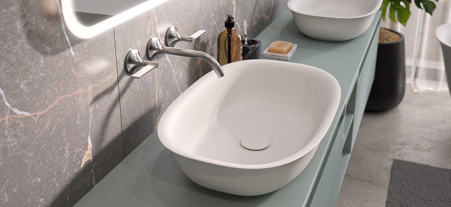 Modernes Badezimmer mit weißem rechteckigen Waschbecken, Wandarmatur, Seifenschale, Flasche, grünem Waschtisch und einer freistehenden Mineralwerkstoffwanne. Marmoraufsatz, Spiegel und Topfpflanze sind teilweise im Hintergrund zu sehen.