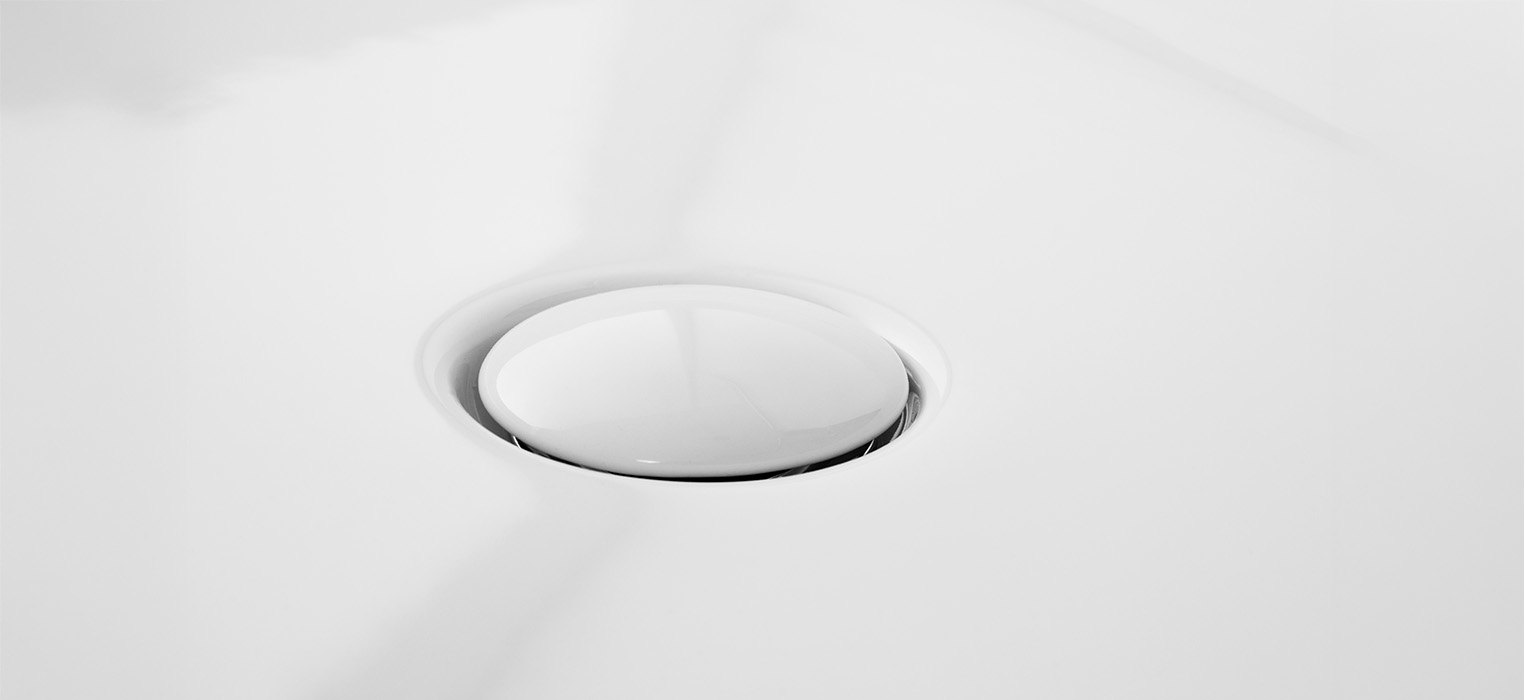 Eine Nahaufnahme eines glänzenden, kreisrunden Abflussstopfens, der in eine glatte, weiße Waschbeckenoberfläche eingelassen ist.