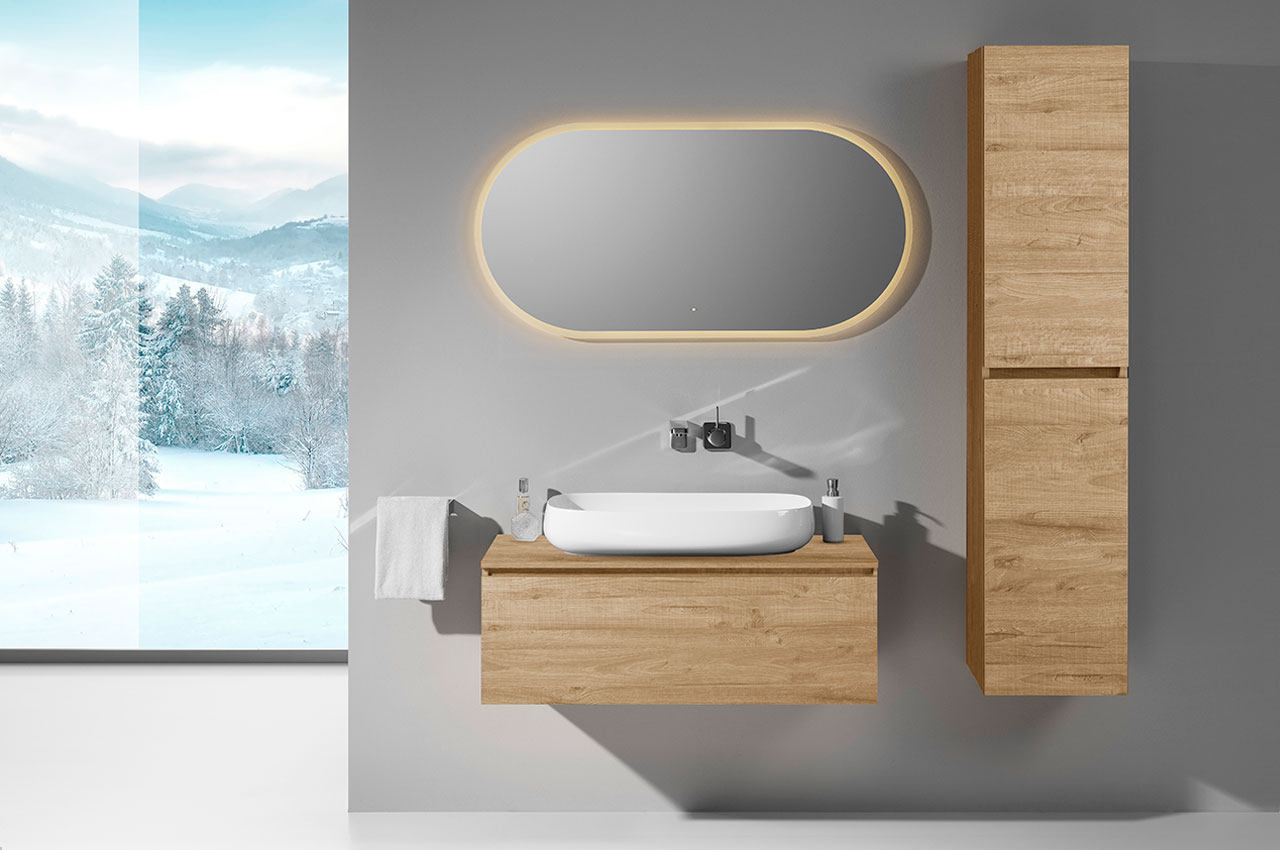 Minimalistisches Bad mit badmöbel holzoptik, einem schwebenden Waschtisch aus hellem Holz, einem großen ovalen Spiegel mit Hintergrundbeleuchtung, einer Wandarmatur, einem hohen passenden Schrank und einer verschneiten Berglandschaft, die durch ein großes Fenster zu sehen ist.