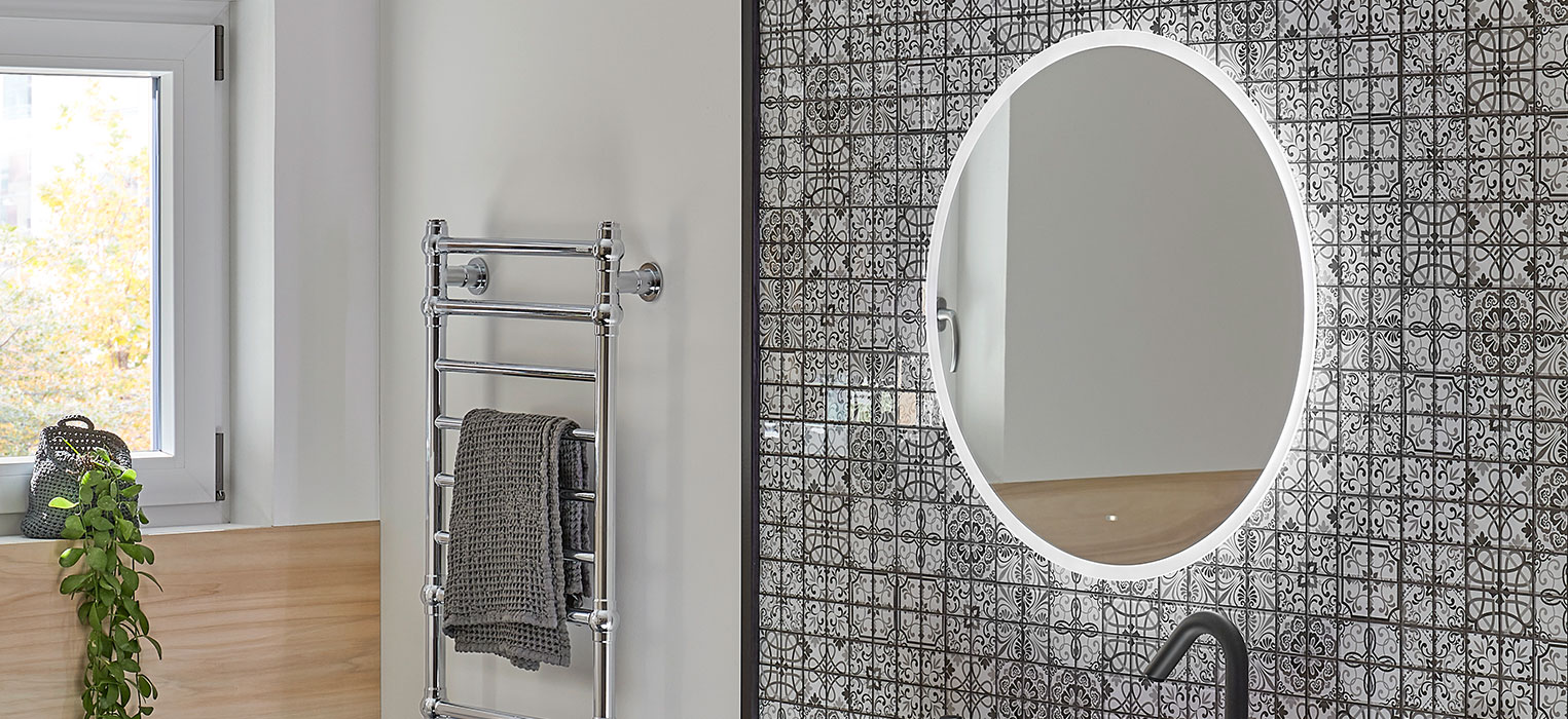 Modernes Badezimmer mit einem runden, hinterleuchteten Spiegel über stilvollen Badmöbeln auf einer gemusterten Fliesenwand, einem verchromten Handtuchhalter mit zwei grauen Handtüchern, einem kleinen Fenster und einer grünen Pflanze auf einem Holzregal.