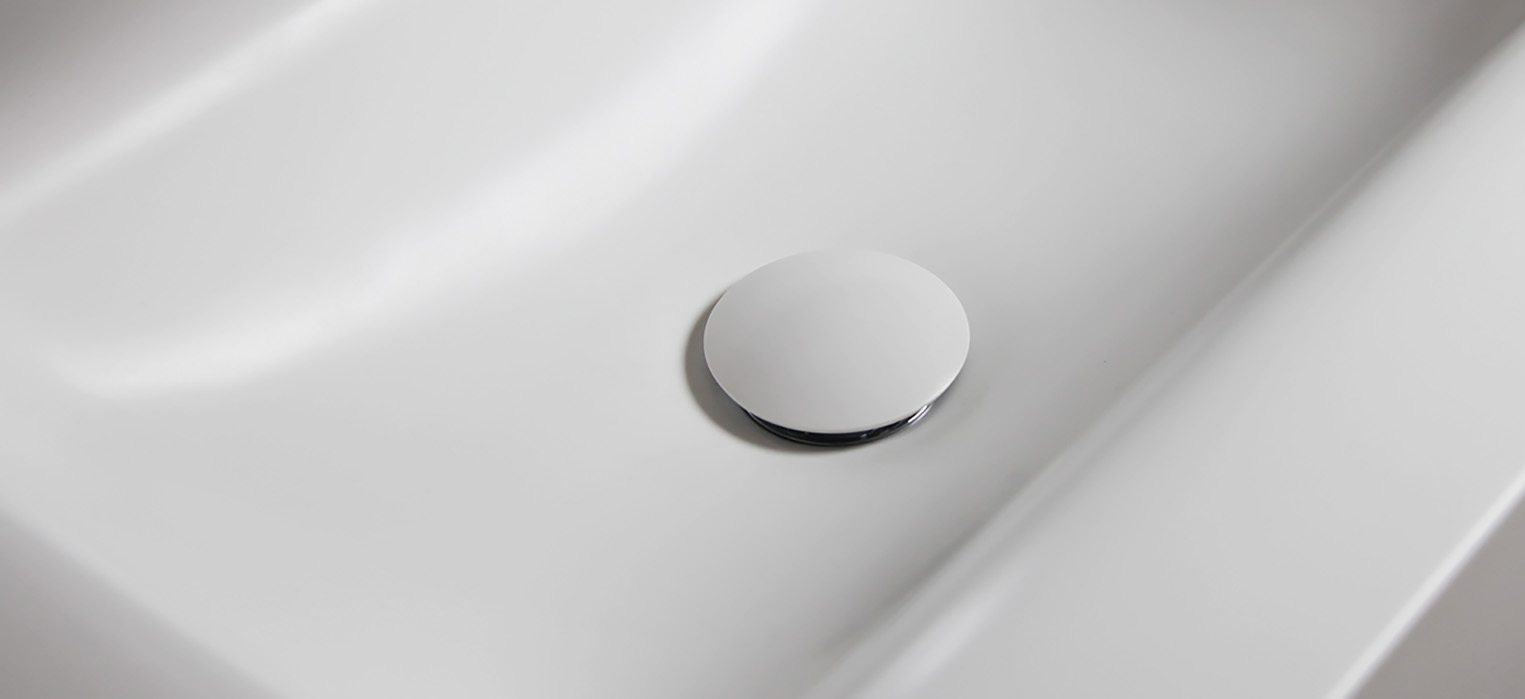 Eine Nahaufnahme eines weißen Waschbeckens mit einer runden, weißen Abflussabdeckung in der Mitte. Die Oberfläche des Waschbeckens ist glatt und sauber und reflektiert das Licht.