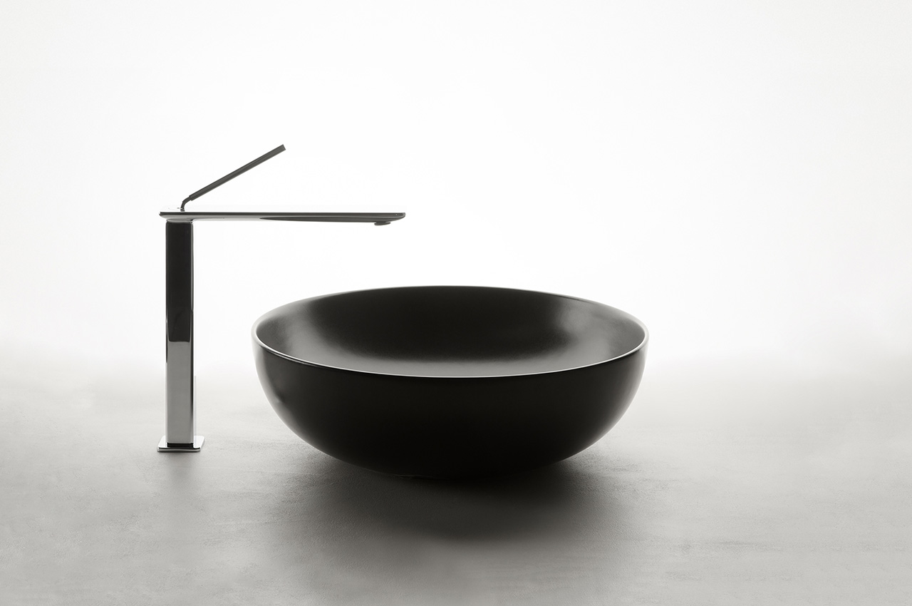 Ein modernes, minimalistisches Waschbecken mit einem runden, mattschwarzen Gefäß und einer schlanken, hohen Chromarmatur auf einer glatten Oberfläche vor einem schlichten weißen Hintergrund.