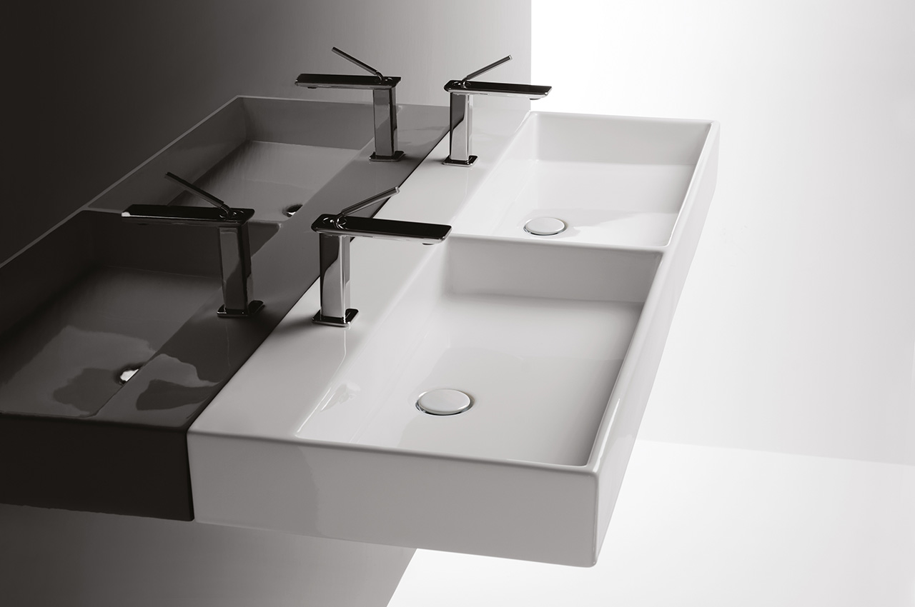 Ein modernes Doppelwaschbecken mit glatten, rechteckigen weißen Becken und minimalistischen Chromarmaturen vor einem zweifarbigen Hintergrund, bei dem eine Seite schattig und die andere hell beleuchtet ist.