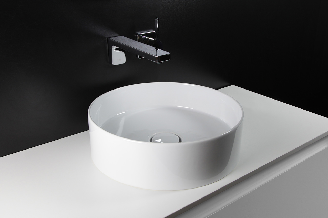 Ein modernes, weißes, rundes Gefäßwaschbecken steht auf einer weißen Arbeitsplatte, hinter der eine schlichte, rechteckige Chromarmatur an einer schwarzen Wand montiert ist. Das gesamte Badezimmer ist minimalistisch und zeitgemäß gestaltet.