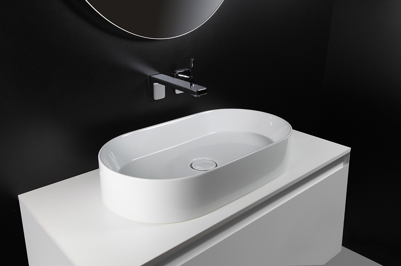 Ein modernes Waschbecken mit einem ovalen weißen Becken, einem verchromten Wasserhahn und einem weißen Waschtisch vor einer schwarzen Wand mit einem runden Spiegel, der am oberen Rand des Bildes teilweise sichtbar ist.