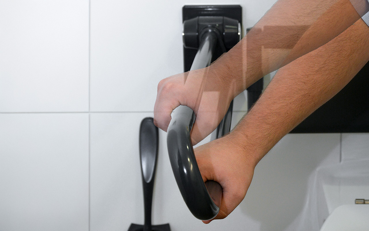 Die Hände einer Person halten eine schwarze Stützstange in einem Badezimmer mit weiß gefliesten Wänden; im Hintergrund sind eine Toilettenbürste und ein Teil einer Toilette zu sehen.