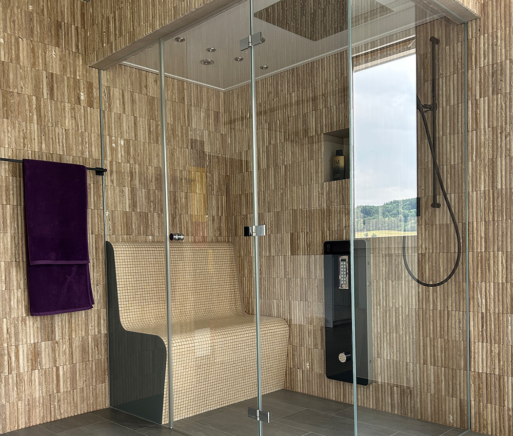 Moderne Dampfdusche mit Glastüren, gefliesten Wänden, einer eingebauten Sitzbank, einem schwarzen Duschpaneel mit Bedienelementen, einer Handbrause und einem violetten Handtuch, das an der linken Wand hängt. Durch das Fenster ist die Aussicht nach draußen zu sehen.