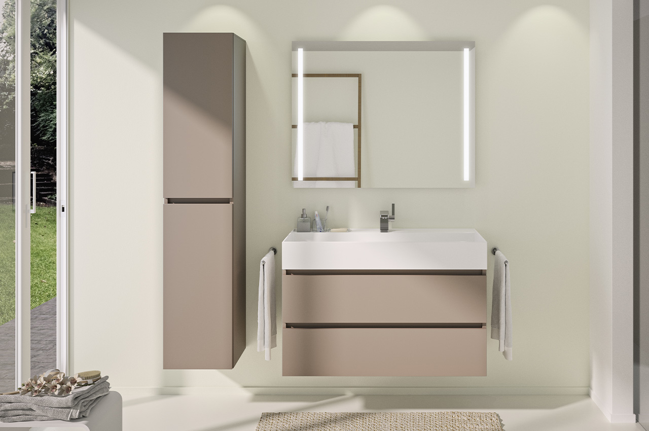 Modernes Badezimmer mit einem beigen, freistehenden Waschtisch, einem großen Spiegel mit integrierter Beleuchtung, einem hohen Schrank, Handtuchhaltern und Sonnenlicht, das durch eine Glastür hereinströmt, die auf einen grünen Garten führt.