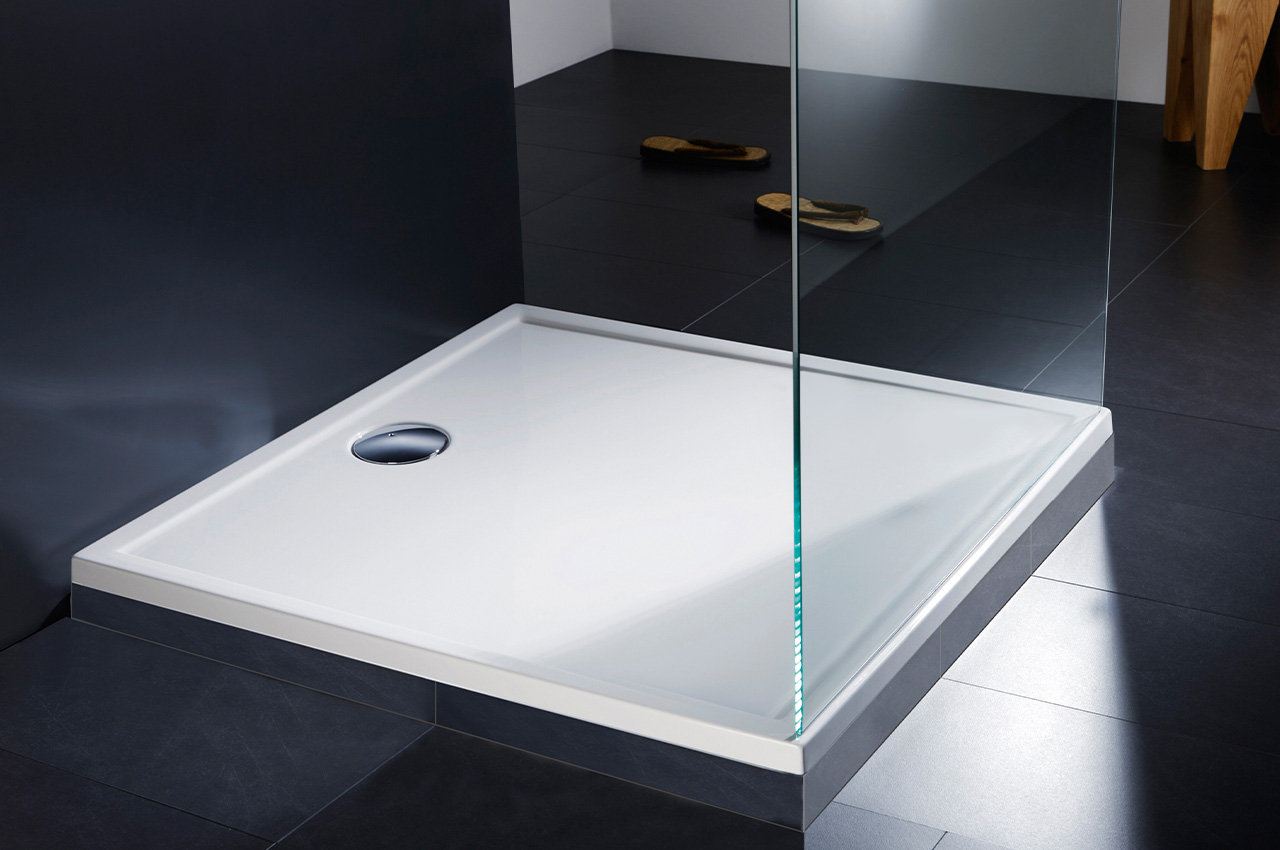 Ein moderner Duschbereich mit einer weißen, quadratischen Duschwanne, einer Glaswand, dunklen Bodenfliesen und einem Paar Sandalen, das außerhalb der Dusche auf dem Boden steht.